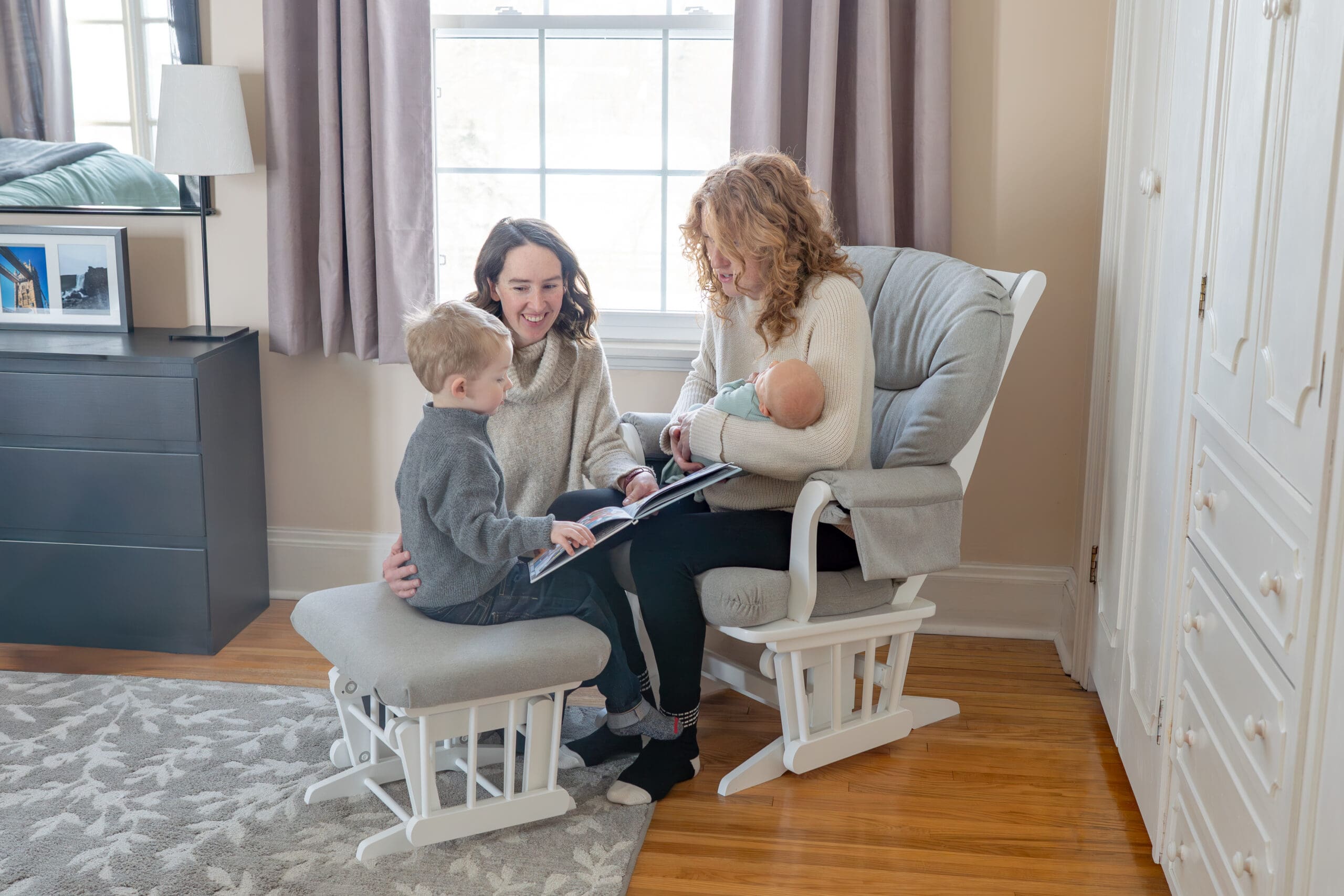Lifestyle newborn session with two moms reading to toddler and holding baby in cozy nursery with natural light — captured by Alliston and Barrie maternity, newborn, and family photographer.