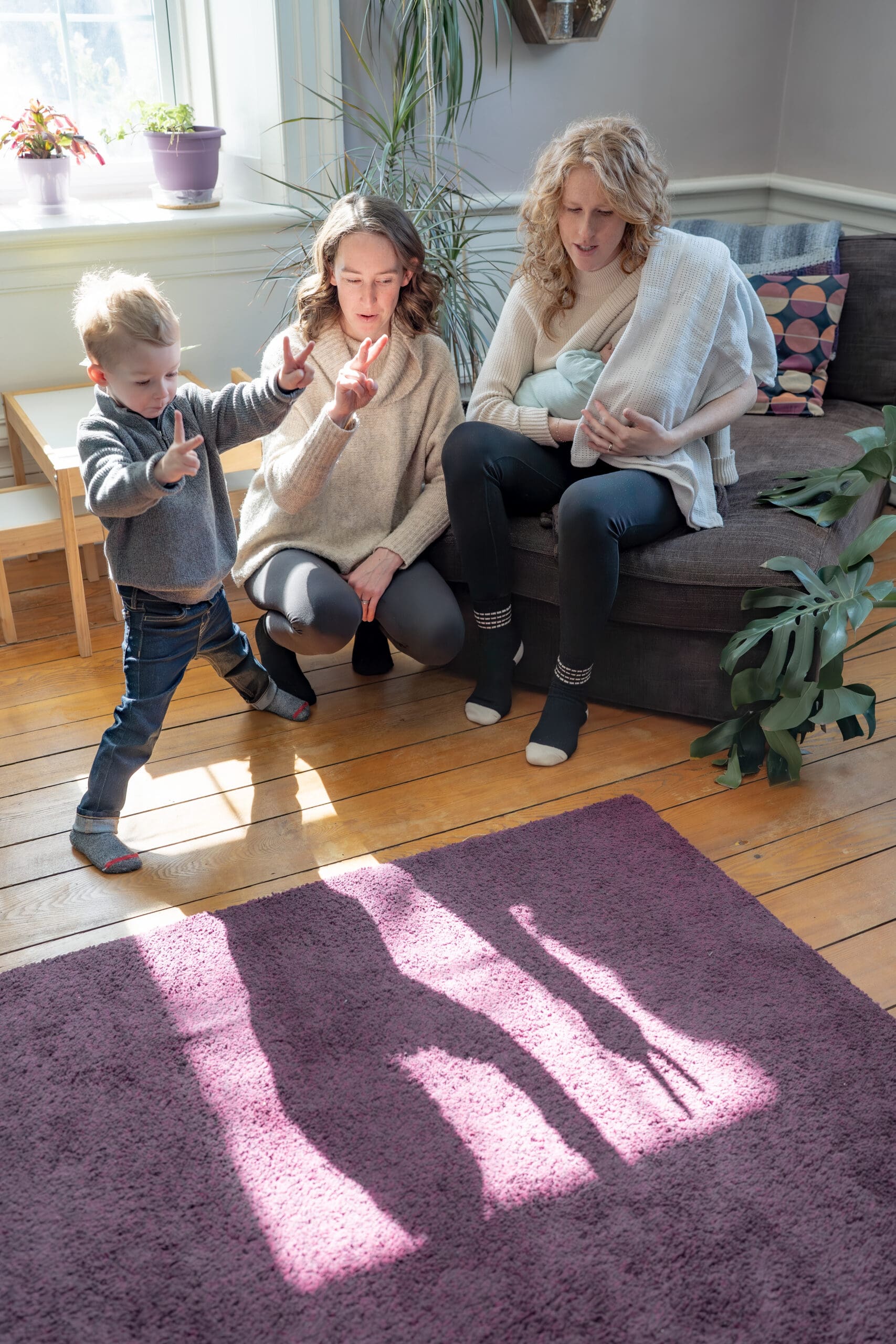 Lifestyle newborn photography session with two mothers sitting in a cozy, sunlit living room with their children—one doing shadow puppets with a toddler, the other holding a newborn baby while sitting on sofa— captured by Alliston and Barrie maternity, newborn, and family photographer.