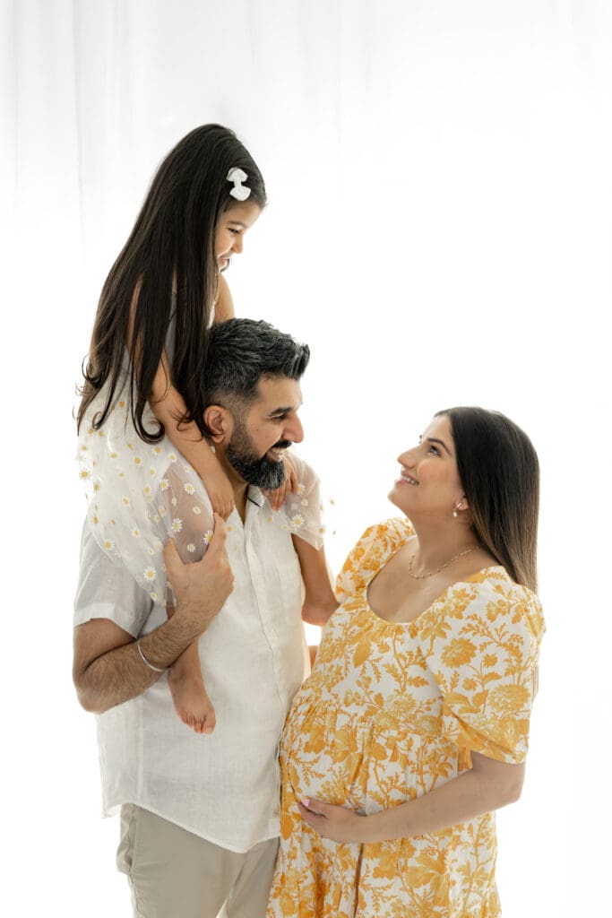 Expecting mother smiling up at toddler sitting on dad's shoulders during a soft, natural light maternity session — photographed by Alliston and Barrie maternity, newborn, and family photographer.
