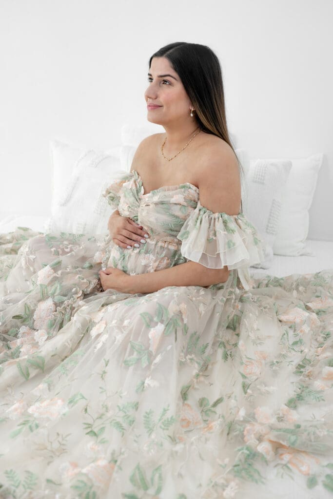 Expecting mother sitting on bed and smiling in floral gown during a soft, natural light maternity session — photographed by Alliston and Barrie maternity, newborn, and family photographer.