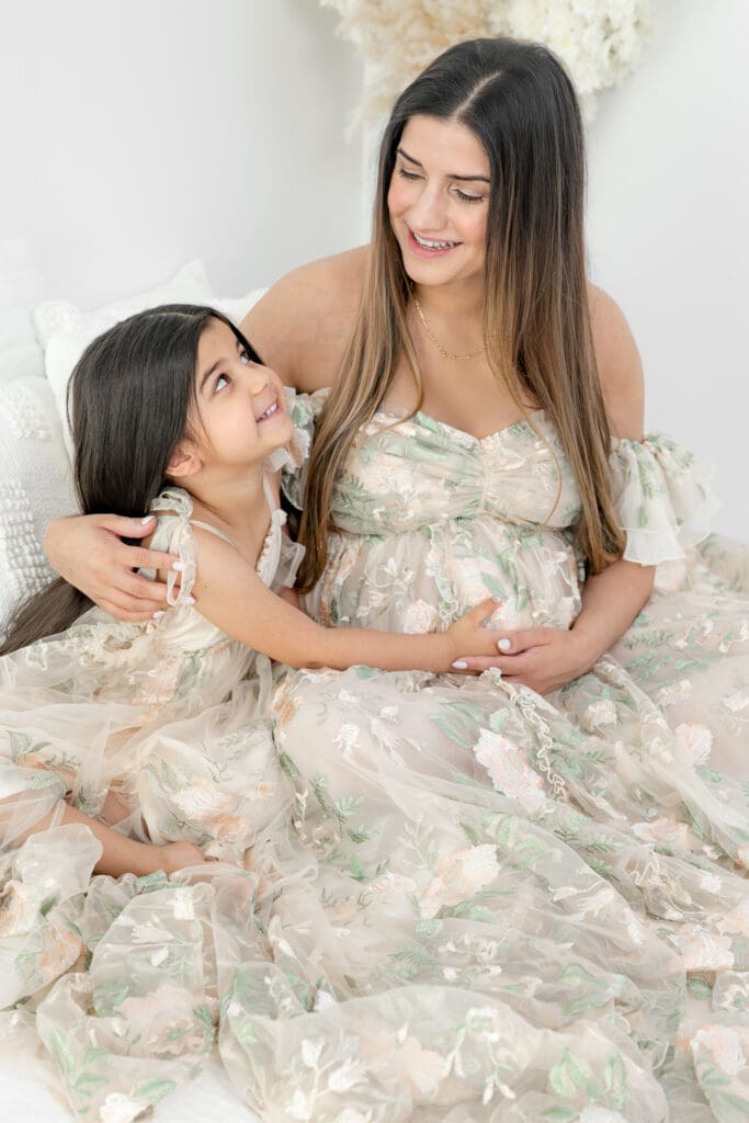 Expecting mother smiling at and hugging toddler in matching floral gowns during a soft, natural light maternity session — photographed by Alliston and Barrie maternity, newborn, and family photographer.