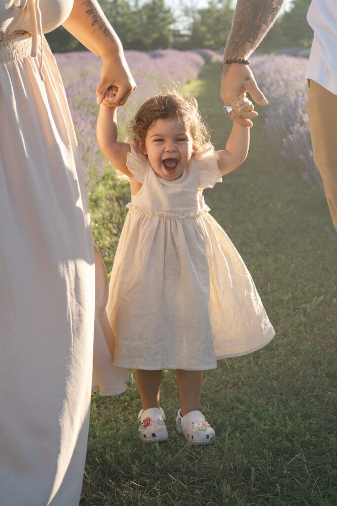 Toddler holding parents' hands and smiling in lavender field during family photography session in Creemore