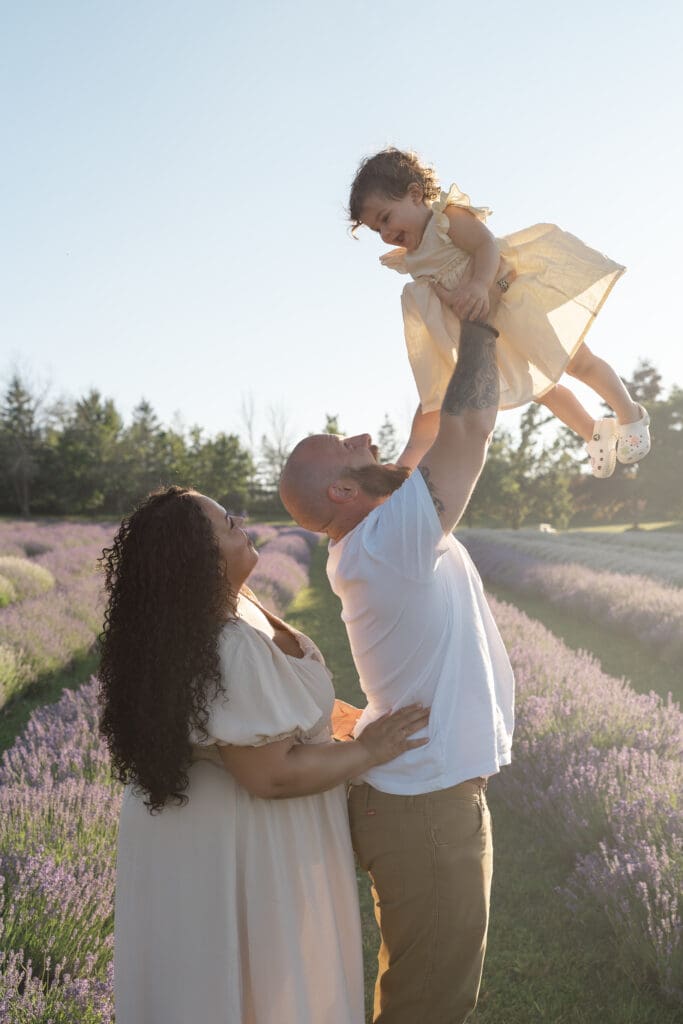 Dad holding toddler up high while mom looks on in lavender field during family photography session in Creemore