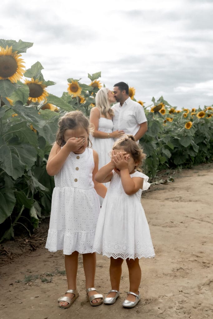 Sisters covering their eyes while parents kiss in sunflower field during family photography session in Innisfil