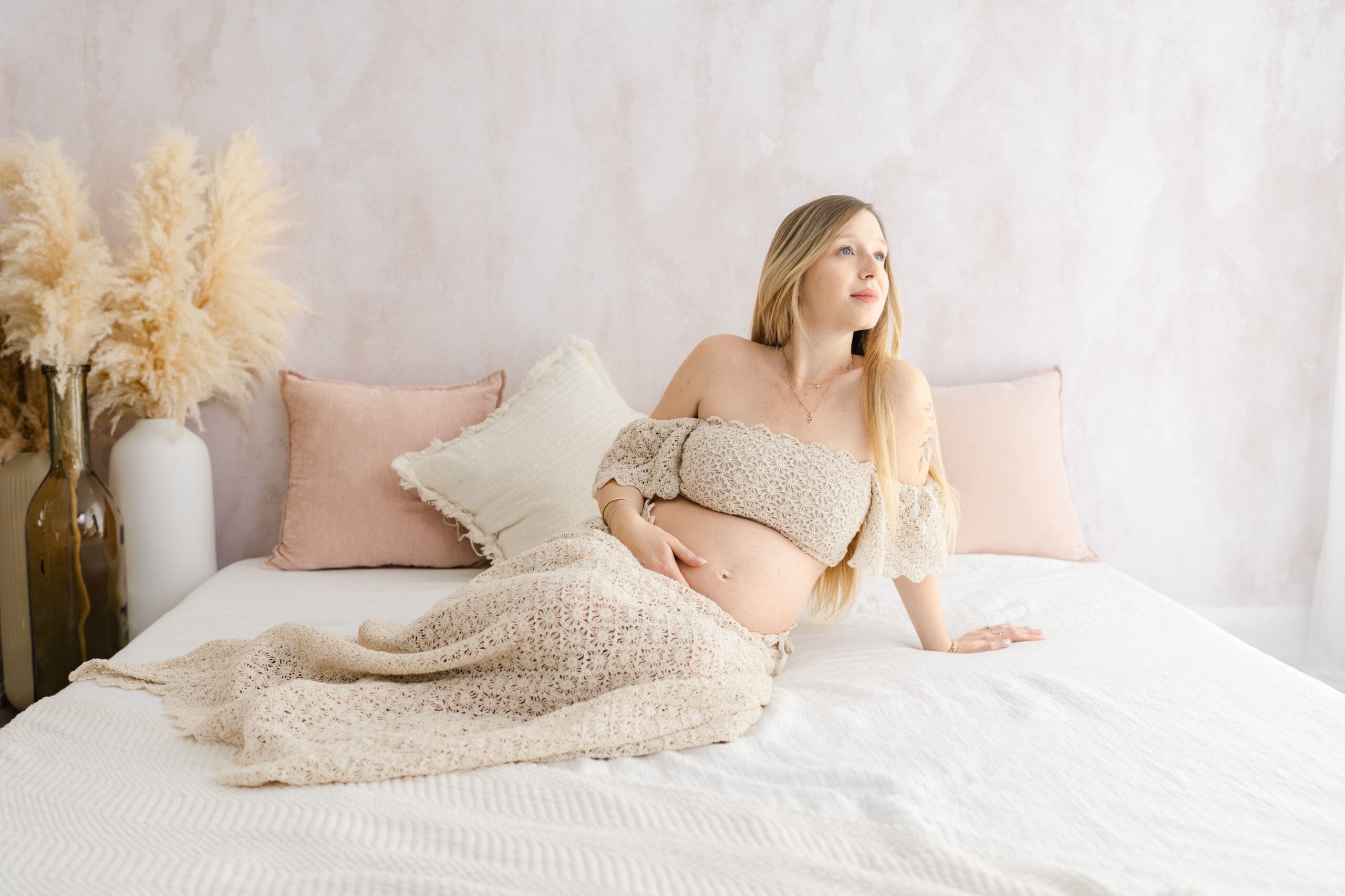 In-studio fine art maternity session in Barrie featuring mom in two-piece gown sitting on bed and holding bump